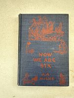 Front cover of the blue cloth hardcover book with orange illustration of a ship and small figures, titled "Now We Are Six" by A.A. Milne.