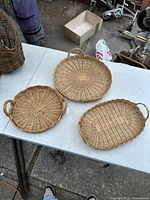 Three handmade woven trays on a white table outdoors, showing different shapes and handles.