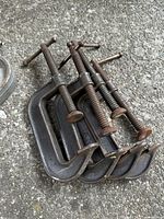 Four 4-inch metal C-clamps, showing wear and rust, stacked together on a concrete surface.