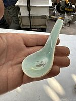 Full view of small celadon spoon in hand showing shape, glaze, and condition including chip.
