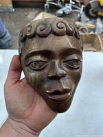 Front view of the hand holding the carved wooden African mask face showing detailed facial features including slightly closed eyes, defined nose, full lips, and spiral curls representing hair.