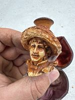 Front view of detailed carved tobacco pipe showing human face with textured hat, held by hand, with open velvet-lined case in background.