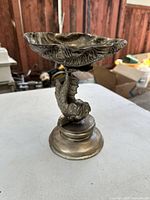 Front view of the antique cast bronze fish-shaped container showing the concave bowl resembling a shell atop a vertically curved fish figure and circular base.