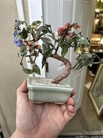Overall front view of antique mini bonsai held in hand showing celadon pot, trunk, jade leaves, and blue and pink jade flowers.