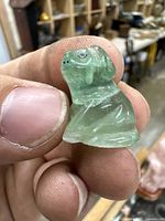 Close-up photo of translucent green carved stone dog netsuke held between fingers, showing detailed carving and facial features
