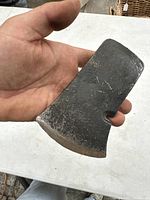 Close-up of an antique hatchet axe head held in a hand showing a blade edge and nail pulling notch, with noticeable wear and rust.