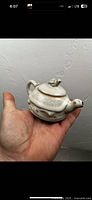 Photo of a small antique Chinese porcelain tea kettle with white celadon glaze held in hand, showing body, handle, lid, and spout with small perforation.