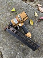 Full side view of the Bostitch framing nailer showing overall shape, yellow body, black magazine, and grip, resting on concrete ground with leaves around it.