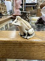 Front and side view of carved frog netsuke showing detailed carving and wear, placed on wood surface with background of other collectibles.