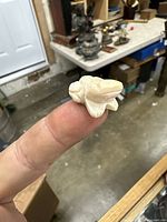 Close-up photo of small carved frog netsuke statue held on a fingertip, showing front and top details with carved features and smooth cream-colored surface.