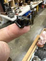 First carved obsidian frog held on fingertip showing glossy dark surface and distinct frog features, photographed in workshop setting.