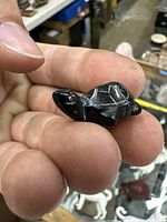 Close-up of miniature turtle carved from black rainbow obsidian held between fingers, showing front and side details, including shell pattern and loop attachment for necklace.