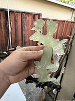 Front view of the pale green jade serpentine carved Guanyin figure held in hand against outdoor background, showing the detailed robes and flower held in hand along with the green wooden base.