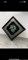 Front view of the framed jade carving displaying the floral pot design with green matte and dark wooden frame set diamond-shaped.