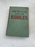 Front cover of the book showing the green cloth cover with title 'One Thousand and One Riddles' in red and black lettering.