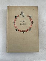 Front cover of the Daniel Boone children's book from 1952 showing beige cloth with red and green floral wreath decoration and a frontiersman illustration.