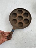 Full view of cast iron aebleskiver pan showing seven round molds and handle with a blue tape mark.