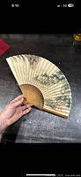 Full view of the folded fan showing the hand-painted paper and bamboo ribs held in hand.