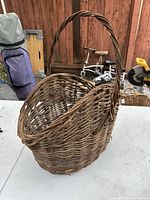 Front and side angle view of a large antique hand woven wicker basket with a tall single handle, placed on a table with a wooden background.