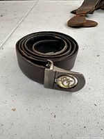 Rolled dark brown leather belt placed on a white surface with a metal buckle showing a circular emblem.