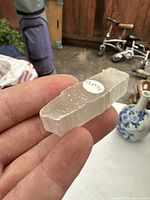 Close-up photo of a small translucent selenite crystal being held between fingers, showing the crystal's natural grain and a round label marked 'Lexite'.