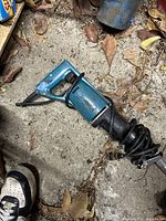 Full length view of Makita Sawzall reciprocating saw lying on ground with coiled power cord attached. Saw shows light wear and dirt.