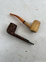 Both antique pipes shown side by side on a light concrete surface, one corn cob pipe and one carved burl wood pipe with bent stems.