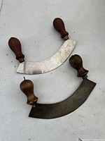 Both antique pizza cutter knives showing crescent-shaped blades and wooden handles, one blade with visible surface rust, the other stamped by maker.