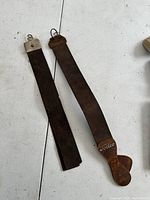 Pair of antique butcher's leather straps laid side by side on a white surface showing straps and metal hardware.