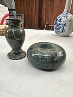 Pair of green granite candleholders on a white surface with a wooden wall background. One candleholder is round and flat with a recessed center, the other is a tall vase shape with a flared opening.
