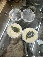 Two carved jade leaf pendants in clear round plastic display cases with white foam interior.