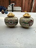 Pair of round Chinese antique salt and pepper shakers with floral cloisonné enamel and gold-tone tops sitting on table.