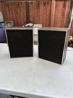 Pair of vintage rectangular speakers with metal frames and black mesh fabric front covers on a white table against a wooden background.