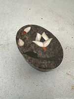 Top and angled view of oval stone box with white and orange flower petals inlaid and black stems on brown stone background.