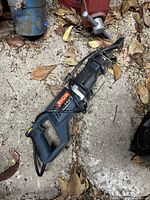 Full view of Ryobi reciprocating saw lying on ground with leaves around, showing saw blade, handle, and body with brand label.