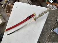 Full view of the katana sword lying on a table showing the blade, handle, and red scabbard.