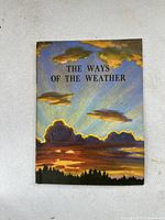 Front cover of 'The Ways of the Weather' textbook showing illustrated cloudy sky with sun rays.