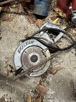 Photo of an old metal Skilsaw circular saw with rust on blade and housing, showing 'SKILSAW' written on side, surrounded by leaves and dirt.