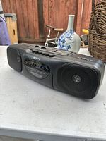 Black vintage Sony boombox placed on white surface outdoors with floral vase and bicycle in background. Shows front view with dual speakers and radio cassette player.