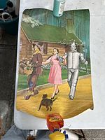 Full view of the Wizard of Oz poster featuring Tin Man, Dorothy, Scarecrow, and Toto on yellow brick road with a log cabin background.