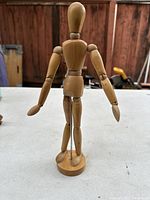 Back view of wooden articulating poseable artist mannequin on round base showing jointed arms, torso, and legs
