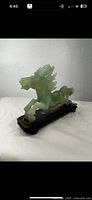 Front-right angled view of light green translucent carved jade horse figurine mounted on a wooden base, showing the horse's detailed mane, head, and stance.