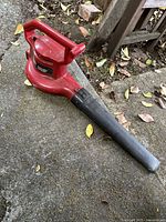 Red corded Toro leaf blower on concrete with leaves around. Shows full side view of blower including handle and nozzle attachment.