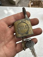 Close-up photo of a heavily aged brass Yale padlock held in hand, showing the rectangular lock body and a key inserted in the bottom keyhole.