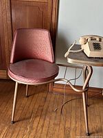 Antique telephone table with an attached chair, showing the pink vinyl cushioned round seat and backrest, metal frame, and wooden top with a telephone on it.