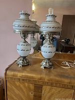 Pair of vintage white glass bedroom lamps with metal bases on wooden surface, reflecting in mirror behind. Both lamps have glass chimney shades and black decorative trim on glass.