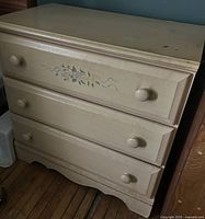 Wooden chest with three drawers, floral design on the top drawer, round wooden knobs, cream color with wear and scuff marks.