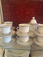 White ceramic coffee cups and matching saucers stacked on a glass surface against a red background. Each cup and saucer has a gray geometric pattern along the edges.