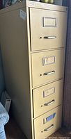 Single beige metal filing cabinet with four drawers shown from an angled front view, located next to a wall and other items.