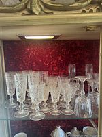 Display cabinet shelf with multiple tall and medium crystal glasses arranged in rows in front of a red textured background.
